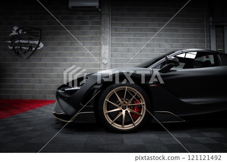 The McLaren 720S is indoors against a gray brick wall with a lion emblem, featuring a matte black finish, gold alloy wheels, and red brake calipers. The McLaren 720S is indoors against a gray brick wall with a lion emblem, featuring a matte black finish, gold alloy wheels, and red brake calipers. 121121492