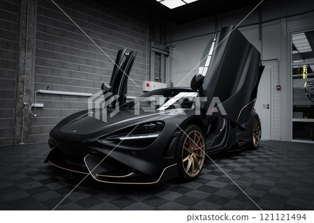 A matte black McLaren 720S with open dihedral doors is set in a modern garage. The car features gold accented wheels and a checkered floor backdrop. 121121494