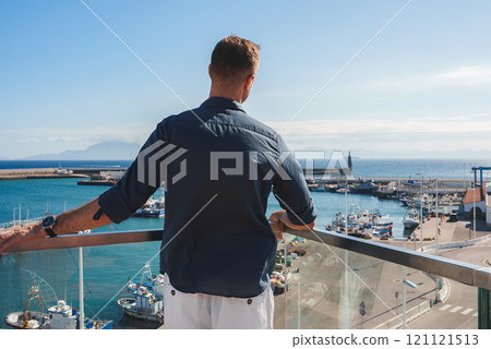A man stands on a balcony overlooking a marina filled with boats, with a lighthouse on a breakwater and distant mountains under a clear blue sky. 121121513