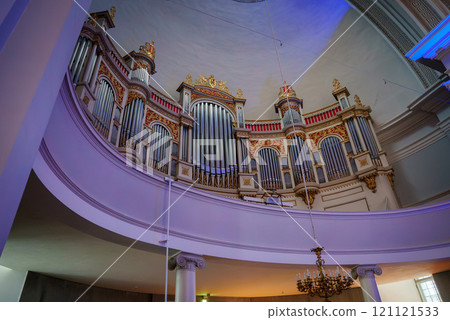 A grand pipe organ with gold and red accents is set against white columns and a curved balcony in a Helsinki church. A chandelier hangs from the domed ceiling. 121121533