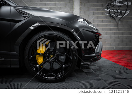 The image shows a close up of a Lamborghini Urus Performante's front wheel with black alloy rims and yellow brake calipers, set against a gray brick wall. 121121546