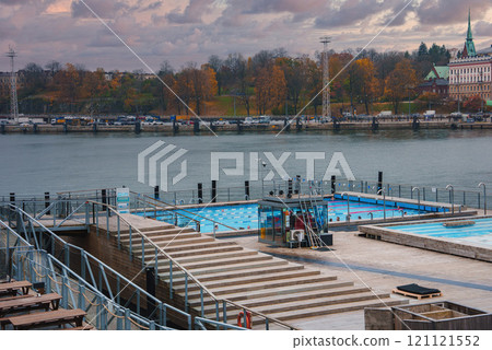 View of Allas Sea Pool by the waterfront in Helsinki, Finland, with modern steps, safety railings, and a cityscape featuring a green spire. 121121552