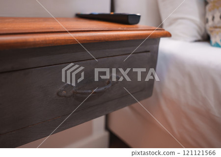 A wooden bedside table with a dark finish and metal handle drawer. A remote control rests on its lighter wood surface. White linens and a pillow are visible. A wooden bedside table with a dark finish and metal handle drawer. A remote control rests on its lighter wood surface. White linens and a pillow are visible. 121121566