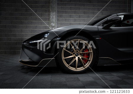 The image features a McLaren 720S with a matte black finish, parked indoors. The focus is on its front section, highlighting bronze wheels and red calipers. 121121633