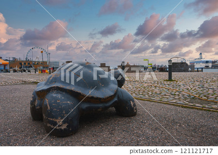 A turtle sculpture stands in a public space in Helsinki, Finland. A ferris wheel and cruise ship are visible in the background, set against an overcast sky. A turtle sculpture stands in a public space in Helsinki, Finland. A ferris wheel and cruise ship are visible in the background, set against an overcast sky. 121121717