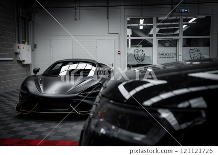 A matte black McLaren 720S is displayed in a well lit workshop with a lion logo on glass doors. A Lamborghini Urus Performante is partially visible. 121121726