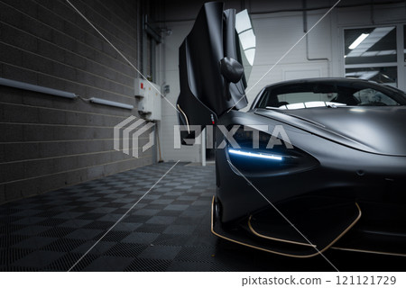 The McLaren 720S is displayed indoors on a checkered floor, featuring its aerodynamic design and LED headlights. A brick wall and piping are visible. 121121729