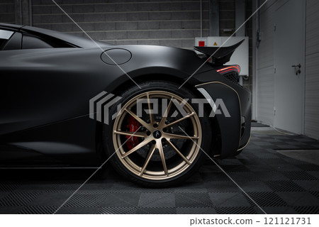 The image captures the rear section of a McLaren 720S, focusing on the gold alloy rim and red brake caliper. The matte black body contrasts with the wheel in an indoor showroom setting. The image captures the rear section of a McLaren 720S, focusing on the gold alloy rim and red brake caliper. The matte black body contrasts with the wheel in an indoor showroom setting. 121121731