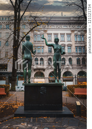 Bronze figures of Zacharias Topelius stand on a pedestal in a Helsinki park. The scene includes ornate architecture, benches, and autumn leaves under an overcast sky. 121121734