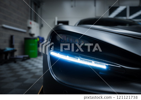 The image captures the illuminated LED headlight of a McLaren 720S indoors, with a tiled floor and industrial elements like a green barrel visible. 121121738