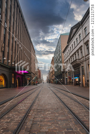 View of a cobblestone street in Helsinki, Finland, featuring tram tracks, historic buildings, and a partly cloudy sky, creating a serene urban scene. 121121758