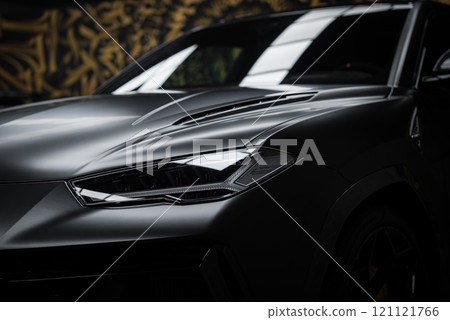 The image showcases the front section of a Lamborghini Urus Performante, highlighting its angular headlights and matte finish, set against a gold and black mural. 121121766