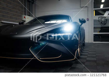 The McLaren 720S is parked indoors on a checkered floor, highlighting its aerodynamic design, LED headlights, and matte black finish in a modern facility. 121121768