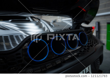 The image shows the rear section of a McLaren 720S, featuring quad exhaust pipes with blue accents. The setting includes a brick wall and industrial elements. 121121783