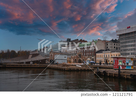 View of Helsinki's waterfront showcasing a red brick building amidst modern structures. The sky's pink and blue hues reflect on the water, creating a vibrant scene. 121121791