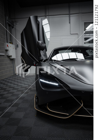 The McLaren 720S is parked indoors on a checkered floor, showcasing its matte black finish with gold accents and distinctive LED headlights. 121121792