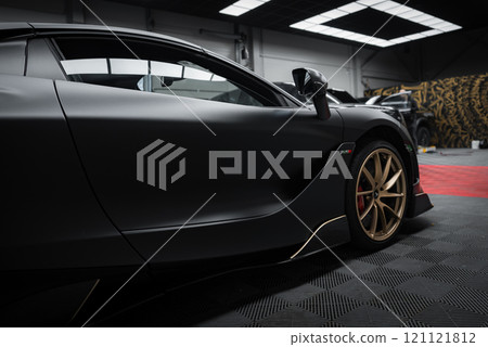 The image features a McLaren 720S indoors, highlighting its sleek design, matte black finish, and gold alloy wheels. A Lamborghini Urus Performante is partially visible in the background. 121121812