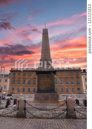 The Keisarinnankivi obelisk stands in Helsinki's Market Square, framed by a vibrant sunset sky. A European style building adds cultural context. 121121813