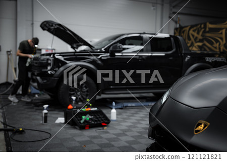 A Lamborghini Urus Performante is parked in a garage with its logo visible. A person works on a black pickup truck in the background, surrounded by tools. 121121821