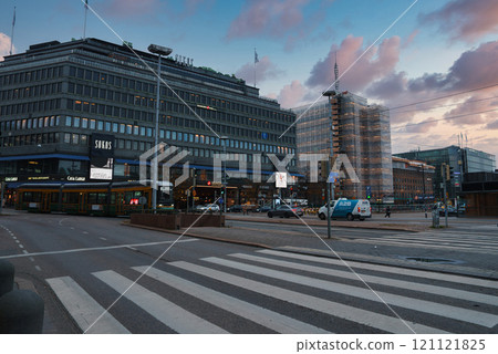 A bustling street in Helsinki city center features the Sokos department store, modern buildings, and a tram. The sky is pink and blue, suggesting dusk. 121121825