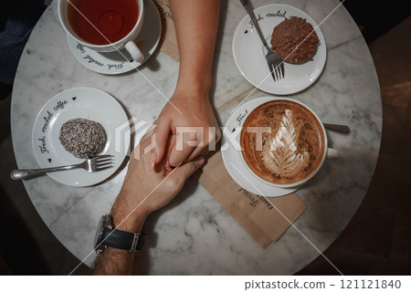 A marble table in a Helsinki cafe features cappuccino with latte art, tea, and desserts. Two hands clasp in the center, creating an intimate scene. A marble table in a Helsinki cafe features cappuccino with latte art, tea, and desserts. Two hands clasp in the center, creating an intimate scene. 121121840