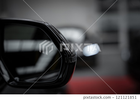 A detailed view of a sleek black side mirror on a luxury supercar, with a blurred background hinting at another vehicle's headlight in a showroom setting. 121121851
