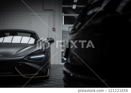 The McLaren 720S is displayed indoors, highlighting its matte black finish, gold accents, and LED headlights. A blurred vehicle adds depth to the scene. 121121856