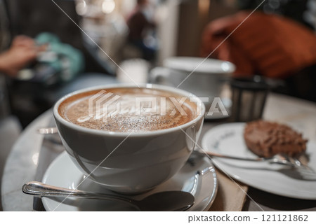 A frothy cappuccino in a white cup on a saucer with a spoon, set in a cozy Helsinki cafe. A blurred dessert and cup in the background add to the ambiance. 121121862