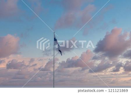 A Finnish flag waves prominently against a serene sky with pastel clouds, showcasing blue and white colors. The scene suggests dawn or dusk in Helsinki. 121121869