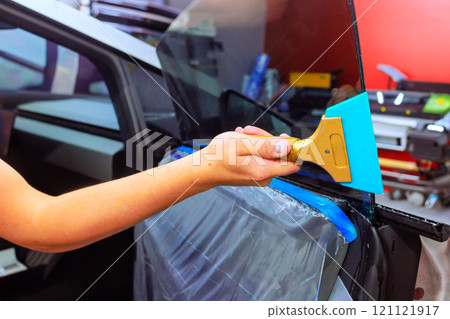 Hand worker carefully applies squeegee to install window film on vehicle side window in workshop environment. 121121917