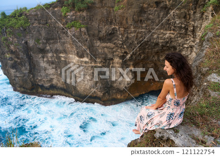 A beautiful woman in a pink dress sits on a cliff above the ocean on the island of Nusa Penida. Devil's Billabong an incredibly wonderful lagoon with splashes from the waves. 121122754