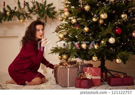 Gorgeous brunette girl quietly puts a box with a gift under the Christmas tree. Holiday surprise for the family 121122757