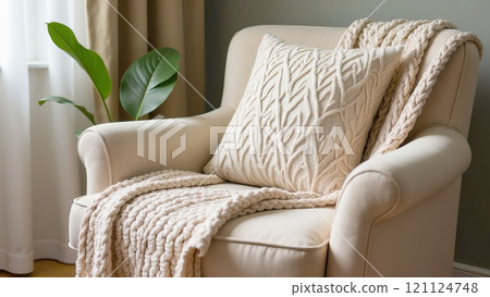 Cosy armchair with thick knitted plaid in dark beige colour and cushion. House decor in rustic style. Still life details in the living room. 121124748