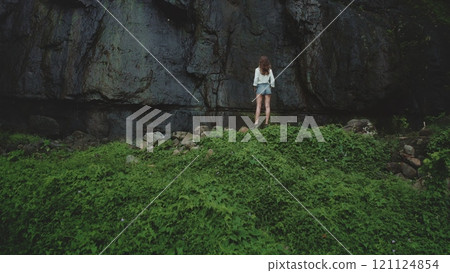 Young woman exploring a tropical island, standing at the edge of lush green vegetation near a towering rock face, feeling tranquil and free in this untouched paradise 121124854