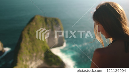 Young woman enjoys a peaceful moment on a cliff, taking in stunning ocean views at sunset on nusa penida island, indonesia 121124904