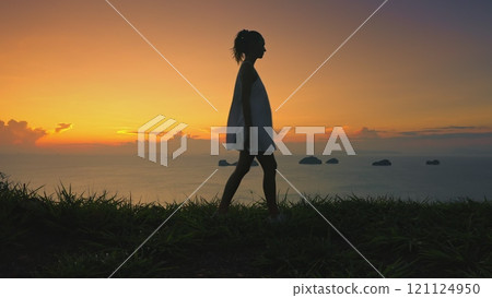 A woman walking on a vibrant green hill sunset sky in background, surrounded by lush grass and rolling landscape. The vast expanse of nature stretches out before her. 121124950