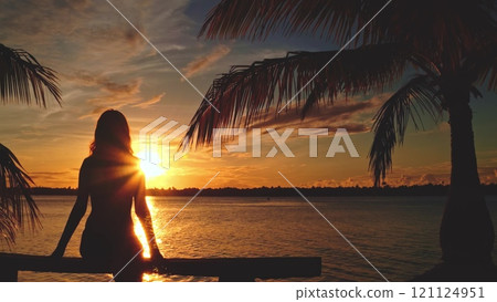 Silhouette of a young woman is sitting on a pier with palm trees at sunset. She is enjoying the view of a tropical lagoon Silhouette of a young woman is sitting on a pier with palm trees at sunset. She is enjoying the view of a tropical lagoon 121124951