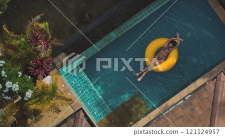 Aerial view of a young woman enjoying her vacation, relaxing on a yellow inflatable ring in a luxurious swimming pool surrounded by palm trees 121124957