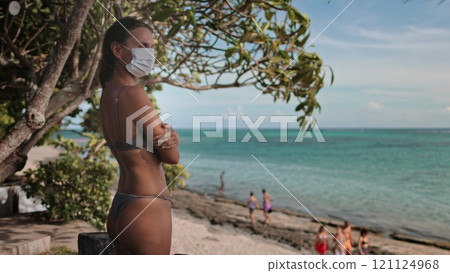 A lone woman wearing a face mask stands on a sandy beach, the vast ocean waves crashing behind her, symbolizing the surreal impact of COVID-19 on summer vacation. 121124968
