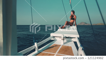 Woman relaxes on yacht sitting on wooden boat nose and looking at ocean wild nature landscape in background. Beautiful slim tanned girl in bikini has travel tour to Indonesia, Komodo National Park. 121124982