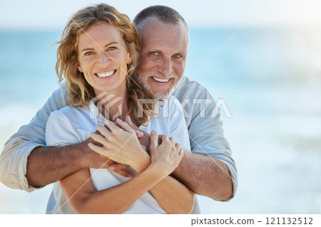 Portrait of happy and loving retired mature caucasian couple enjoying a romantic date at the beach on a sunny day. Cheerful affectionate husband hugging his wife while enjoying seaside vacation 121132512