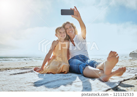 Carefree mother and daughter taking a selfie while sitting on the beach. Happy little girl and grandmother smiling while taking a picture on a cellphone while on holiday. Mom and daughter bonding 121132533