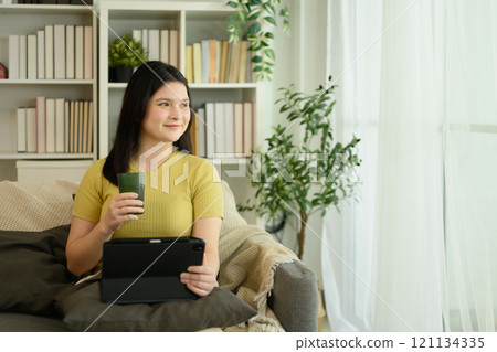 Smiling young woman relaxing on sofa, holding a green mug and using digital tablet 121134335