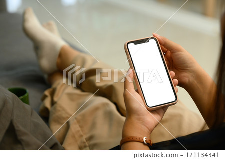 Close up of woman hands holding a smartphone with white screen sitting on sofa 121134341