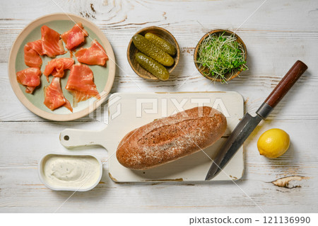 Top view of ingredients for a sandwich - salmon, pickles, watercress sprouts, a loaf of bread, a knife, and a lemon 121136990