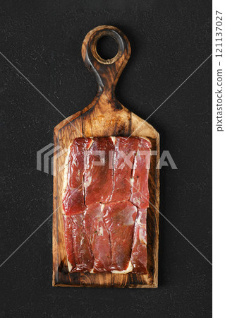 Block of cured duck meat on a handcrafted wooden cutting board against a dark background 121137027