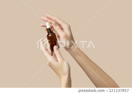 A womans hands holding a brown bottle of serum on a beige background. Body care 121137657