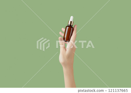 A womans hand holding a brown bottle of serum on a green background 121137665