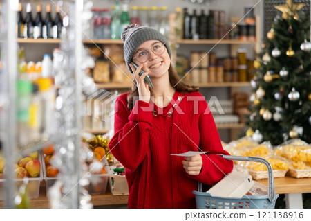 She holds paper note in hands, asks mother questions on phone while shopping in grocery 121138916
