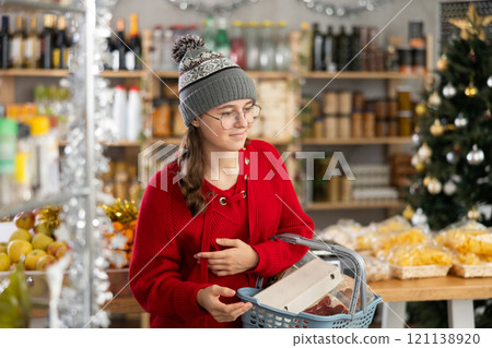 Female shopper with grocery cart attentively choosing fresh food in grocery supermarket for Christmas celebration 121138920